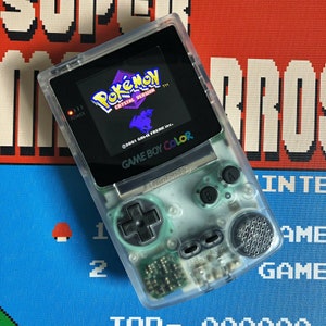 以下が含まれることがあります： 透明なGame Boy Colorのゲーム機で、Pokémon Crystal Versionのゲームが表示されています。画面にはゲームのタイトルと紫色のシルエットが表示されています。コンソールには方向パッド、AボタンとBボタン、スピーカーがあります。「GAME BOY COLOR」の文字が見えます。