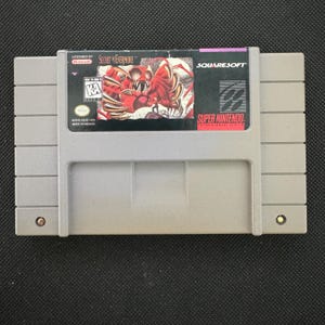 Puede incluir: Un cartucho de videojuego gris de Super Nintendo para el juego Secret of Evermore de Squaresoft. La etiqueta presenta una ilustración de un monstruo rojo, el sello oficial de Nintendo, una clasificación KA y el logo de Super Nintendo.