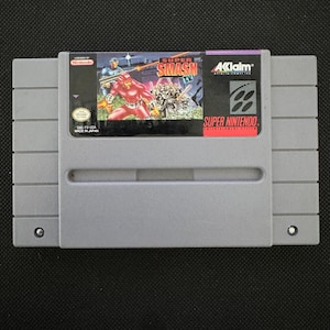 Puede incluir: Un cartucho de videojuego gris de Super Nintendo para Super Smash T.V. La etiqueta muestra a un personaje vestido de rojo sosteniendo un arma, el título del juego en texto rojo y blanco, el logotipo de Acclaim y el sello oficial de Nintendo.