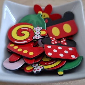 May include: A white square dish filled with colourful cartoon character-shaped magnets. The magnets feature red, yellow, black, and white designs, including Mickey and Minnie Mouse, and other whimsical shapes. The dish sits on a light brown wooden surface.