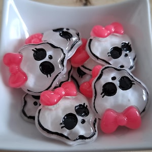 May include: A selection of white skull-shaped beads with black facial features and pink bows, displayed in a white square dish. The beads have a glossy finish and are on a wooden surface.