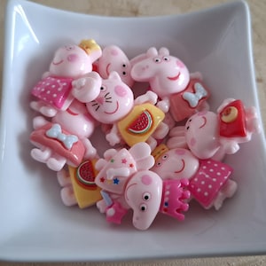 May include: A white square dish filled with pink pig-shaped charms. The charms feature different outfits and accessories, such as dresses, bows, and watermelon slices. The plastic charms have smiling faces and are brightly coloured.