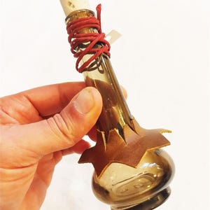 Puede incluir: Una pequeña botella de vidrio marrón con un tapón de corcho y un cordón rojo alrededor del cuello. La botella tiene una pieza decorativa de cuero marrón en la base. Una pequeña etiqueta está unida al cordón.