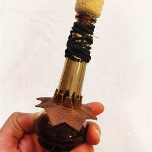 May include: A small, brown glass bottle with a cork stopper. The bottle is wrapped with black cord and has a brown leather starburst design at the base. The bottle is held in a hand against a white background.