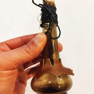 Könnte beinhalten: Eine braune Glasflasche mit Korken und schwarzen Schnurdetails. Die Flasche hat eine braune Lederverzierung am Boden. Die Flasche wird in einer Hand gehalten.