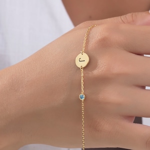 Könnte beinhalten: Ein goldenes Kettenarmband mit einem kleinen blauen Edelstein und einem runden Anhänger, auf dem der Buchstabe "J" eingraviert ist.