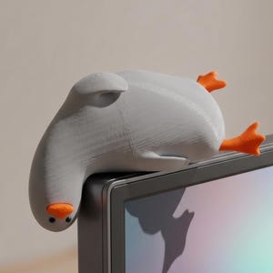 May include: A gray goose-shaped desk accessory with orange feet and beak, perched on the corner of a computer monitor. The goose is made of a textured material and has a whimsical design. The monitor displays a colorful, iridescent screen.