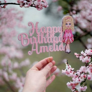Puede incluir: Topper de pastel de purpurina rosa con el texto "Happy Birthday Amelia" y una ilustración de dibujos animados de una niña con un vestido rosa y una falda verde.
