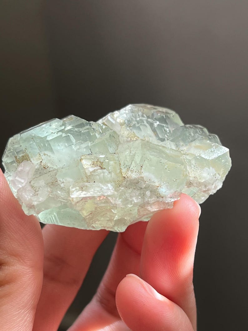 Puede incluir: Un racimo de cristales de fluorita transl&uacute;cidos de color verde p&aacute;lido con una estructura c&uacute;bica. El esp&eacute;cimen mineral presenta bordes geom&eacute;tricos afilados e inclusiones internas visibles, mostrando un tono verde menta claro en toda la formaci&oacute;n.