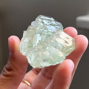 Puede incluir: Un racimo de cristales minerales transl&uacute;cidos de color verde p&aacute;lido con una estructura geom&eacute;trica facetada. El esp&eacute;cimen muestra un tono menta claro y una superficie v&iacute;trea y reflectante con m&uacute;ltiples puntas entrelazadas.