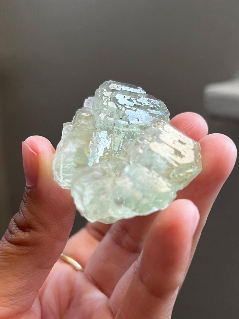 Puede incluir: Un racimo de cristales minerales transl&uacute;cidos de color verde p&aacute;lido con una estructura geom&eacute;trica facetada. El esp&eacute;cimen muestra un tono menta claro y una superficie v&iacute;trea y reflectante con m&uacute;ltiples puntas entrelazadas.