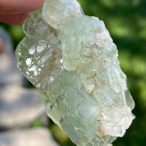 Puede incluir: Un esp&eacute;cimen mineral transl&uacute;cido de color verde p&aacute;lido que presenta un grupo de formaciones cristalinas angulares y bloqueadas. La superficie muestra una mezcla de facetas lisas y v&iacute;treas con inclusiones blancas opacas y texturizadas.