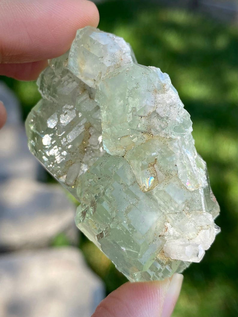 Puede incluir: Un esp&eacute;cimen mineral transl&uacute;cido de color verde p&aacute;lido que presenta un grupo de formaciones cristalinas angulares y bloqueadas. La superficie muestra una mezcla de facetas lisas y v&iacute;treas con inclusiones blancas opacas y texturizadas.