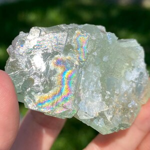 Puede incluir: Un esp&eacute;cimen de cristal de fluorita verde p&aacute;lido transl&uacute;cido con una estructura cristalina irregular. La superficie presenta fracturas internas distintivas que crean vibrantes refracciones de luz arco&iacute;ris en las facetas del mineral.