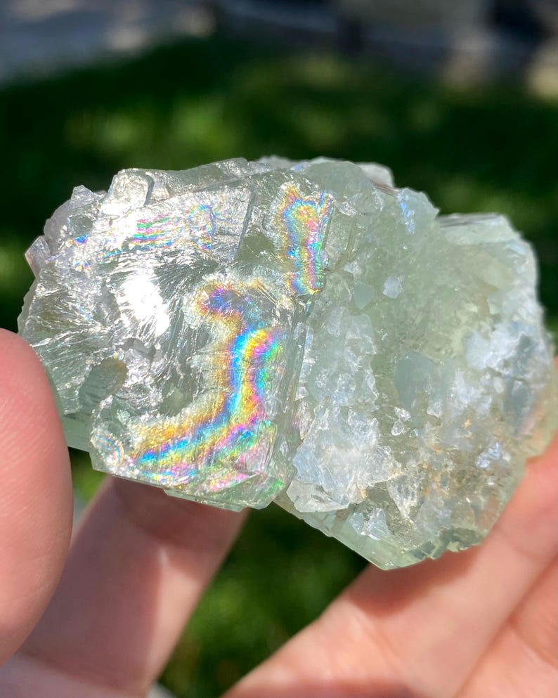 Puede incluir: Un esp&eacute;cimen de cristal de fluorita verde p&aacute;lido transl&uacute;cido con una estructura cristalina irregular. La superficie presenta fracturas internas distintivas que crean vibrantes refracciones de luz arco&iacute;ris en las facetas del mineral.
