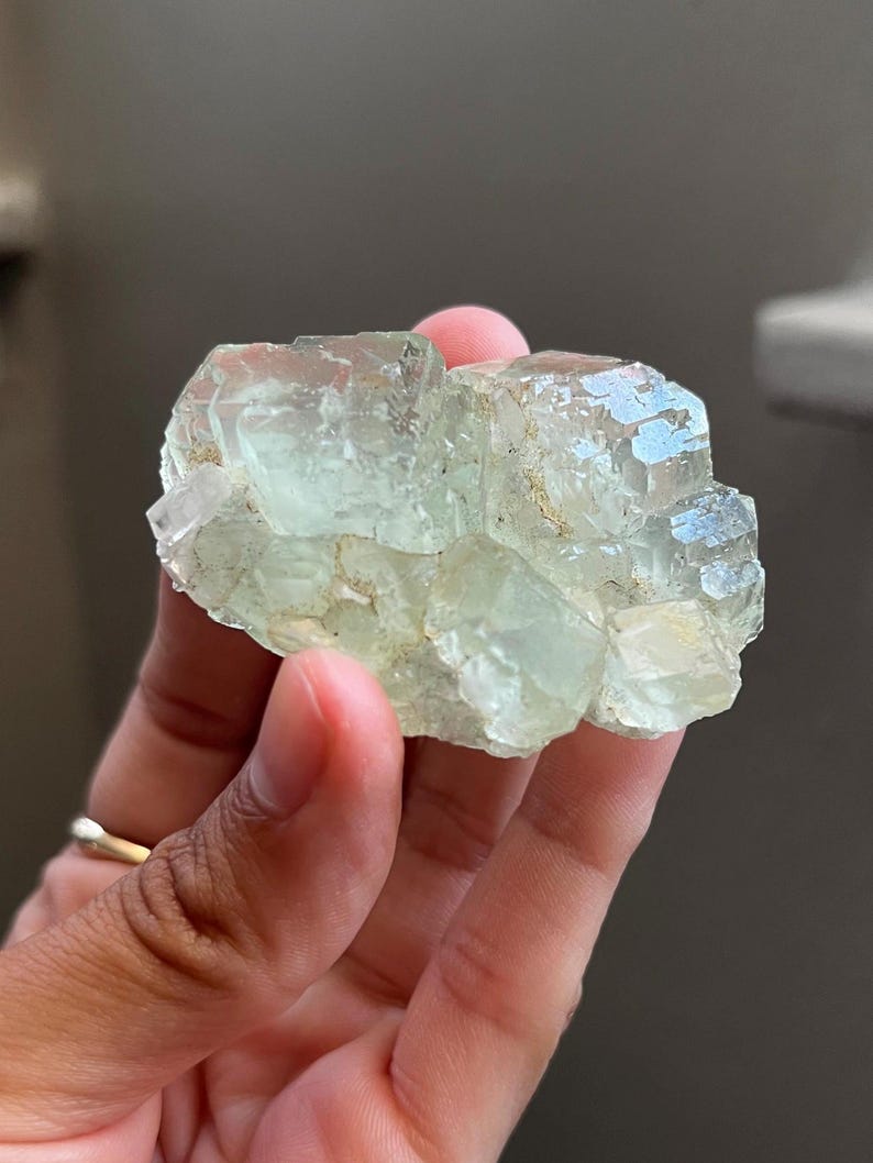 Puede incluir: Un grupo de cristales de fluorita transl&uacute;cidos de color verde p&aacute;lido con estructura c&uacute;bica. El esp&eacute;cimen mineral presenta bordes geom&eacute;tricos afilados y una superficie v&iacute;trea y reflectante con sutiles inclusiones internas y dep&oacute;sitos minerales de color claro.