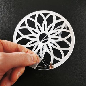 Puede incluir: Una plantilla circular decorativa blanca con un diseño de mandala. La plantilla se está despegando de un respaldo de plástico transparente.