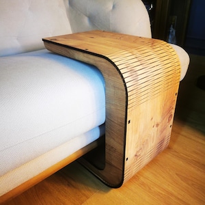 May include: A wooden sofa arm table with a curved design. The table is made of light-coloured wood with a black trim and is designed to fit over the arm of a sofa. The table is shown on a white sofa.