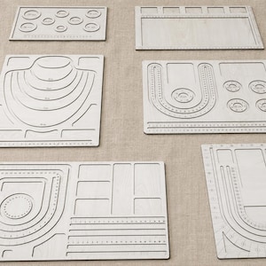 Puede incluir: Una colección de seis tableros de diseño de joyería de madera clara que presentan varios canales grabados, recortes circulares y marcas de medición para organizar cuentas y collares durante el proceso de creación de joyas.