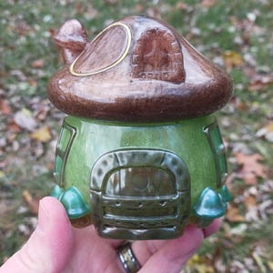 May include: A whimsical ceramic mushroom house with a brown cap and green base. The house features a detailed door, windows, and decorative accents. The mushroom house is a charming decorative item.
