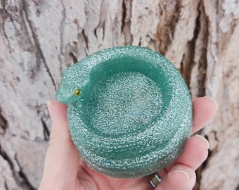 May include: A teal-colored, glittery snake-shaped trinket dish. The coiled snake has yellow eyes and a circular opening for holding small items. The dish is held in a hand against a blurred, textured background.