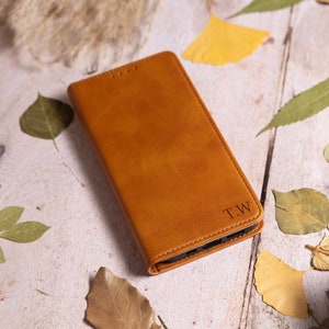 Może przedstawiać: Brązowe skórzane etui na telefon z wygrawerowanymi inicjałami "T.W." na tylnej stronie. Etui leży na drewnianej powierzchni, wokół której rozrzucone są suche liście.