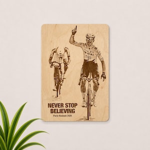Peut inclure: Plaque en bois représentant deux cyclistes, l'un célébrant une victoire. Le texte "NEVER STOP BELIEVING" et "Paris-Roubaix 2026" sont imprimés en dessous. L'image est en nuances de marron.