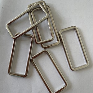 May include: A group of six silver metal rectangular rings, stacked on top of each other. The rings are smooth and shiny.