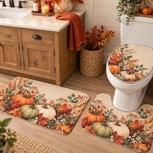 Op de afbeelding: Een driedelige badkamerset bestaande uit badmatten en een toiletdekselhoes met een print van oranje, groene en witte pompoenen, omringd door herfstbladeren, tarwearen en kleine bessen op een lichtbeige achtergrond met een textuur.
