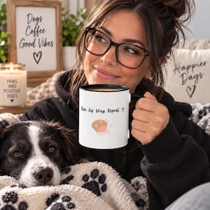 Könnte beinhalten: Ein weißer Keramikbecher mit schwarzem Rand und Henkel, der den schwarzen Text Rise. Sip. Wag. Repeat. neben einem kleinen Kaffeetassensymbol und einer beigen Pfotenabdruckgrafik zeigt. Der Becher wird in der Hand gehalten, nahe einem schwarz-weißen Hund.