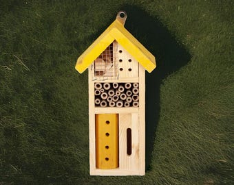 May include: A wooden bee house with a yellow roof and accents. The house features multiple compartments, including a section with bamboo tubes and a yellow block with holes. A metal loop is attached for hanging.