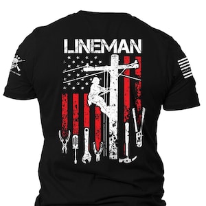 May include: Black t-shirt with a white graphic of a lineman working on a power line in front of an American flag with tools. The text "LINEMAN" is at the top of the graphic.