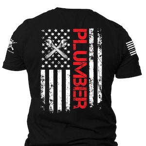 May include: Black t-shirt with a distressed American flag design. The flag is white and red with a red vertical stripe that says "PLUMBER" in white letters. The flag is centered on the back of the shirt. There are two crossed wrenches in the top left corner of the flag.