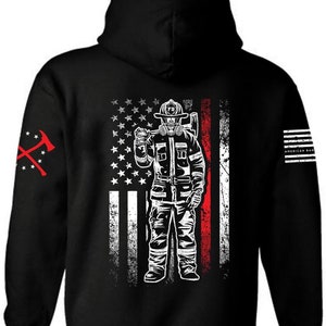 Pu&ograve; includere: Felpa con cappuccio nera con una grafica bianca di un vigile del fuoco in piedi davanti a una bandiera americana con una striscia rossa. Il vigile del fuoco indossa un casco e una maschera antigas. La manica sinistra ha una grafica rossa di due asce incrociate.