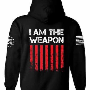 May include: Black hooded sweatshirt with the text "I AM THE WEAPON" in white with a red and white American flag design. The left sleeve has a skull and crossbones with the text "ND" and stars.