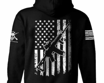 May include: Black hooded sweatshirt featuring a distressed American flag design with a silhouette of a rifle on the back. The left sleeve has a skull and crossbones graphic with the text "Dedicated to the Core". The right sleeve has the text "American Badass" in a distressed font.