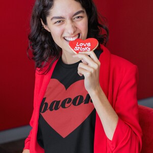 May include: A woman wearing a black t-shirt with a red heart and the word "bacon" printed on it. She is holding a red heart-shaped cookie that says "Love Stinks".