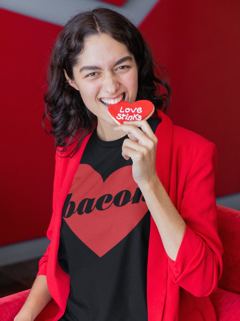 May include: A woman wearing a black t-shirt with a red heart and the word "bacon" printed on it. She is holding a red heart-shaped cookie that says "Love Stinks".