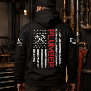 May include: A black hooded sweatshirt featuring a distressed American flag graphic on the back. The design includes two crossed wrenches and the word PLUMBER in red vertical text. A small graphic with the text LIFE PLUMBING is on the right sleeve.