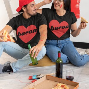 May include: A couple wearing matching black t-shirts with a red heart and the word "bacon" printed on them. They are sitting on the floor with a pizza box, wine, and beer. The woman is holding a slice of pizza and the man is holding a bottle of beer.