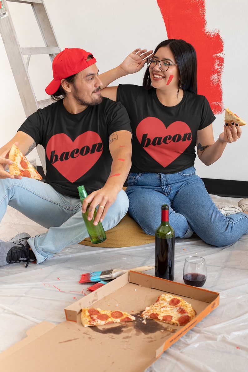 May include: A couple wearing matching black t-shirts with a red heart and the word "bacon" printed on them. They are sitting on the floor with a pizza box, wine, and beer. The woman is holding a slice of pizza and the man is holding a bottle of beer.