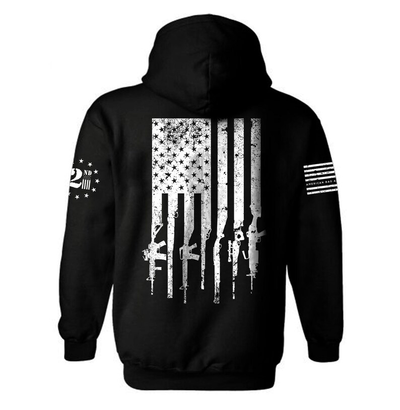 Puede incluir: Sudadera con capucha negra con un dise&ntilde;o de bandera estadounidense blanca desgastada en la espalda. La bandera tiene una fila de rifles delante. La manga izquierda tiene el texto "2nd Amendment" con estrellas alrededor. La manga derecha tiene el texto "American Bad A**" con una peque&ntilde;a bandera estadounidense.