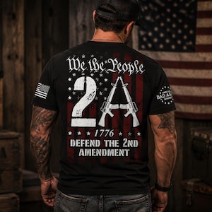 May include: A black t-shirt featuring a graphic on the back with the text We the People, 2A, 1776, and Defend the 2nd Amendment. The design includes two rifles forming the letter A, set against a faded American flag background in red and white.