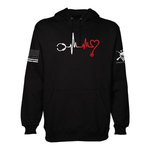 May include: Black hooded sweatshirt with a white heartbeat graphic and a red heart. The graphic includes a stethoscope on each side of the heartbeat. The left sleeve features a faded American flag with the text "PATRIOTIC" on the bottom. The right sleeve features a skull and crossbones with the text "TO THE CORE".