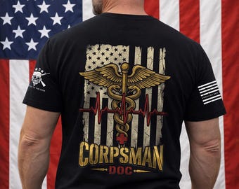 May include: A black t-shirt featuring a graphic on the back with a distressed American flag, a gold caduceus symbol, a red EKG heart rate line, and a red cross. The text reads CORPSMAN and DOC. A small logo is on the sleeve and chest area.