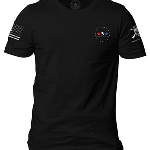 Pode incluir: Camiseta preta com um c&iacute;rculo branco no peito que apresenta tr&ecirc;s estrelas, duas vermelhas e uma azul, com o texto "AMERICAN MADE & SOLD" dentro do c&iacute;rculo. A manga direita tem uma caveira branca e ossos cruzados com o texto "PATRIOTIK" abaixo.