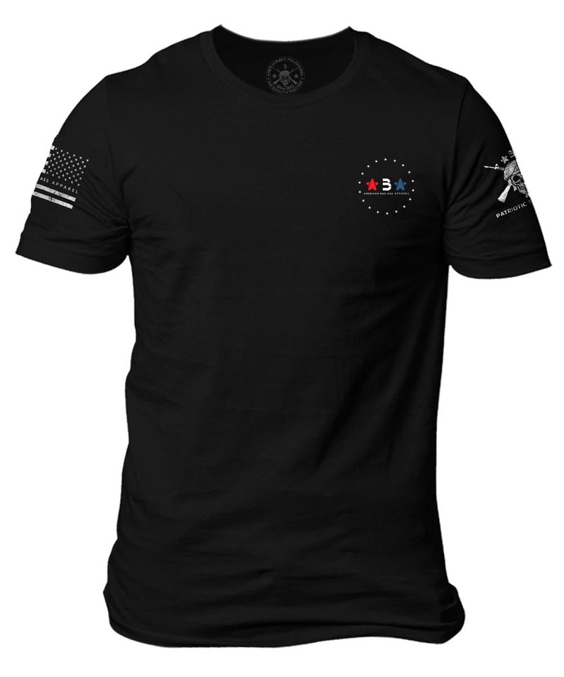 Pode incluir: Camiseta preta com um c&iacute;rculo branco no peito que apresenta tr&ecirc;s estrelas, duas vermelhas e uma azul, com o texto "AMERICAN MADE & SOLD" dentro do c&iacute;rculo. A manga direita tem uma caveira branca e ossos cruzados com o texto "PATRIOTIK" abaixo.
