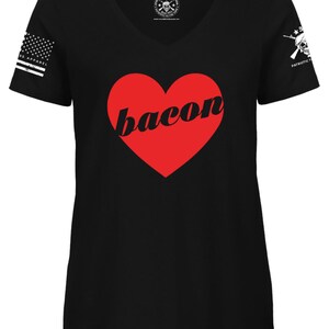 May include: Black V-neck t-shirt with a red heart that says "bacon" in a white font. The shirt has a white American flag on the left sleeve and a skull with crossed rifles on the right sleeve.