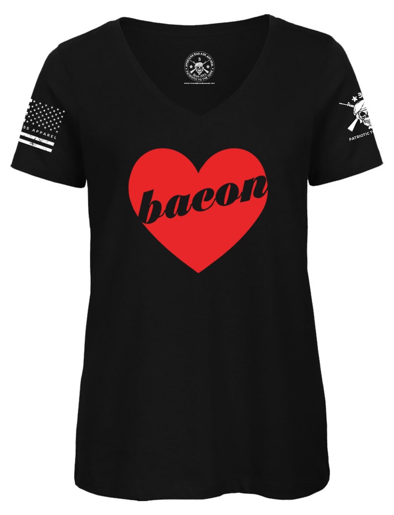 May include: Black V-neck t-shirt with a red heart that says "bacon" in a white font. The shirt has a white American flag on the left sleeve and a skull with crossed rifles on the right sleeve.