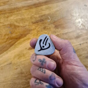 May include: A grey guitar pick with a black design of a stylised hand with three fingers extended. The pick is being held by a hand with tattoos.